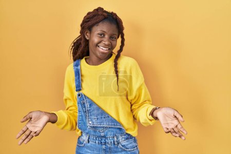 Téléchargez les photos : Femme africaine debout sur fond jaune souriant gai avec les bras ouverts comme accueil amical, salutations positives et confiantes - en image libre de droit
