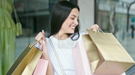 Photo for Young beautiful hispanic woman smiling going shopping holding bags at clothing store - Royalty Free Image