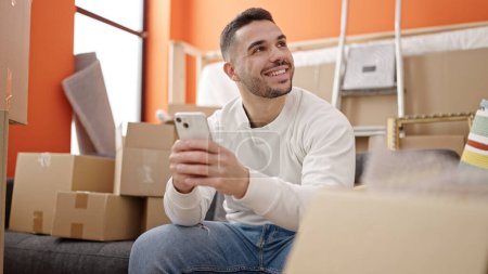 Photo for Young hispanic man using smartphone sitting on sofa at new home - Royalty Free Image