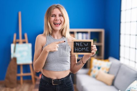 Téléchargez les photos : Blonde femme caucasienne tenant tableau noir avec nouveau texte à la maison souriant heureux pointant avec la main et le doigt - en image libre de droit