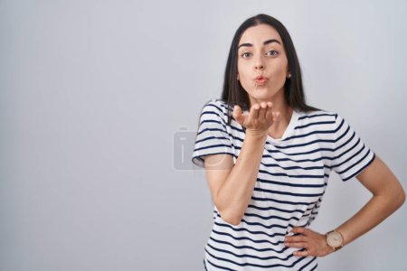 Téléchargez les photos : Jeune femme brune portant t-shirt rayé regardant la caméra souffler un baiser avec la main sur l'air étant belle et sexy. expression d'amour. - en image libre de droit