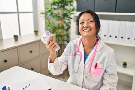 Photo for Young chinese woman wearing doctor uniform using thermometer at clinic - Royalty Free Image