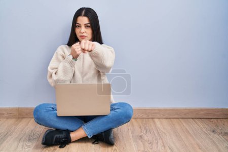 Téléchargez les photos : Jeune femme utilisant un ordinateur portable assis sur le sol à la maison poing de frappe pour se battre, agression agressive et en colère, menace et violence - en image libre de droit