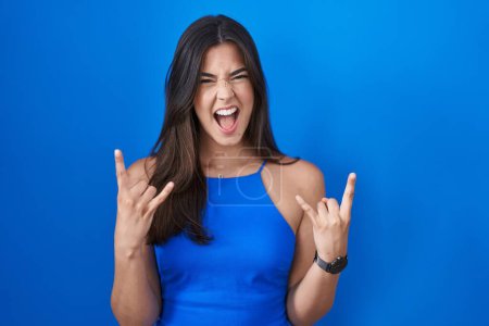 Foto de Mujer hispana de pie sobre fondo azul gritando con expresión loca haciendo símbolo de roca con las manos arriba. Estrella de la música. concepto de música pesada. - Imagen libre de derechos