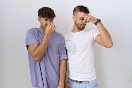 Photo for Homosexual gay couple standing over white background smelling something stinky and disgusting, intolerable smell, holding breath with fingers on nose. bad smell - Royalty Free Image