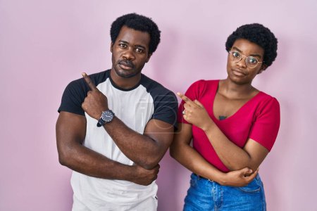 Téléchargez les photos : Jeune couple afro-américain debout sur fond rose pointant avec le doigt de la main sur le côté montrant la publicité, visage sérieux et calme - en image libre de droit