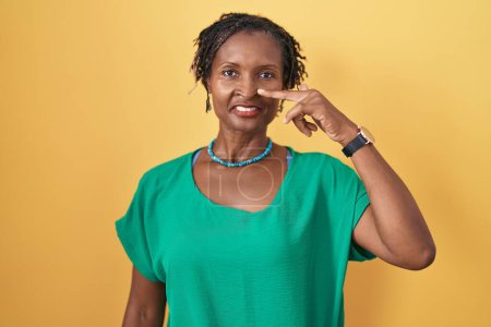 Téléchargez les photos : Femme africaine avec dreadlocks debout sur fond jaune pointant avec le doigt de la main vers le visage et le nez, souriant gai. concept de beauté - en image libre de droit