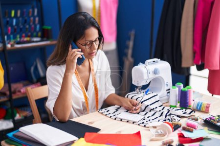 Téléchargez les photos : Jeune femme hispanique tailleur parler sur smartphone écriture sur ordinateur portable au studio de couture - en image libre de droit