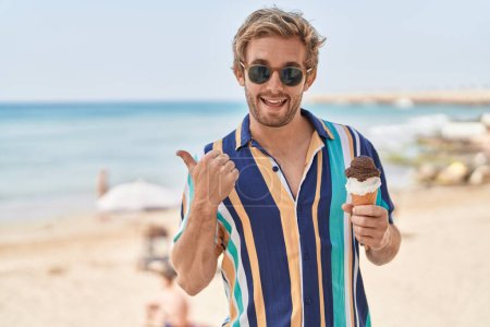 Photo for Caucasian man eating an ice cream at the beach pointing thumb up to the side smiling happy with open mouth - Royalty Free Image