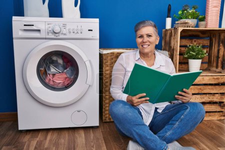 Photo for Middle age woman reading book waiting for washing machine at laundry room - Royalty Free Image