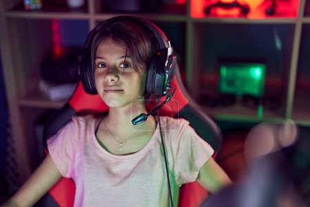 Photo for Adorable hispanic girl streamer sitting on table with serious expression at gaming room - Royalty Free Image