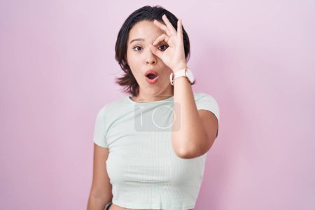 Téléchargez les photos : Jeune femme hispanique debout sur fond rose faisant un geste correct choqué par le visage surpris, les yeux regardant à travers les doigts. expression incrédule. - en image libre de droit