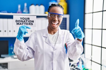 Téléchargez les photos : Belle femme noire travaillant au laboratoire scientifique tenant votre bannière de questions de don souriant heureux et positif, pouce levé faire excellent et signe d'approbation - en image libre de droit