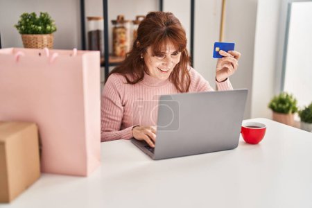 Téléchargez les photos : Femme d'âge moyen achetant en utilisant une carte de crédit et un ordinateur portable assis sur la table à la maison - en image libre de droit