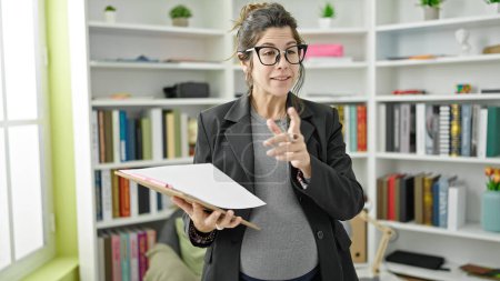 Téléchargez les photos : Jeune enseignante enceinte tenant un presse-papiers expliquant la leçon à la bibliothèque universitaire - en image libre de droit