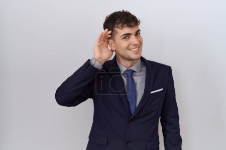 Téléchargez les photos : Jeune homme d'affaires hispanique portant costume et cravate souriant avec la main sur l'oreille en écoutant une rumeur ou des commérages. concept de surdité. - en image libre de droit