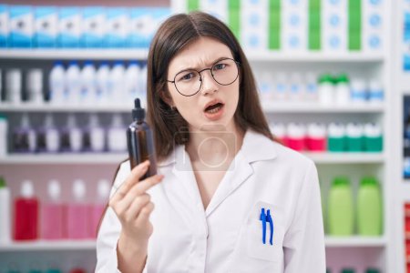 Photo for Young caucasian woman working at pharmacy drugstore holding serum scared and amazed with open mouth for surprise, disbelief face - Royalty Free Image