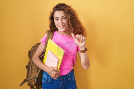 Téléchargez les photos : Jeune femme caucasienne portant un sac à dos étudiant et tenant des livres pointant vers le dos avec la main et les pouces levés, souriant confiant - en image libre de droit
