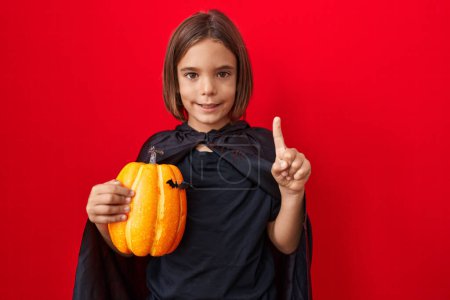 Foto de Pequeño niño hispano usando una capa y sosteniendo la calabaza de Halloween señalando el pulgar hacia el lado sonriendo feliz con la boca abierta - Imagen libre de derechos