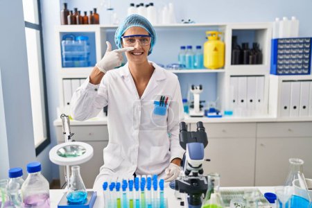 Téléchargez les photos : Femme brune travaillant au laboratoire scientifique pointant du doigt et du nez, souriante et joyeuse. concept de beauté - en image libre de droit