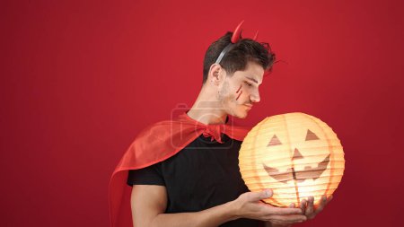 Foto de Joven hombre hispano con traje de diablo sosteniendo lámpara de calabaza de halloween sobre fondo rojo aislado - Imagen libre de derechos