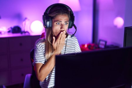 Foto de Joven rubia jugando videojuegos cubriendo la boca con la mano, conmocionada y temerosa de equivocarse. expresión sorprendida - Imagen libre de derechos
