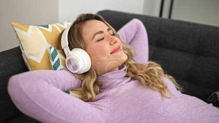 Young blonde woman listening to music lying on sofa at home