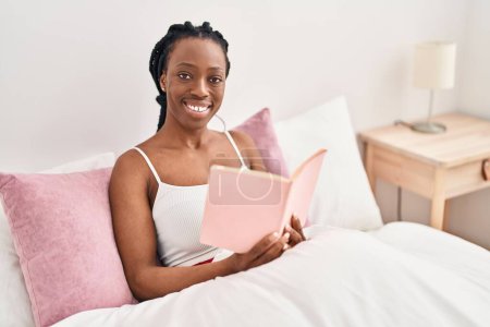 Téléchargez les photos : African american woman reading book sitting on bed at bedroom - en image libre de droit
