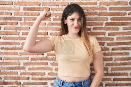 Téléchargez les photos : Jeune femme brune debout sur le mur de briques personne forte montrant muscle du bras, confiant et fier de la puissance - en image libre de droit