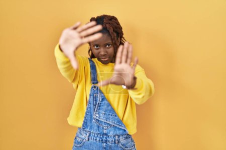 Foto de Mujer africana de pie sobre fondo amarillo haciendo marco usando las manos palmas y los dedos, perspectiva de la cámara - Imagen libre de derechos