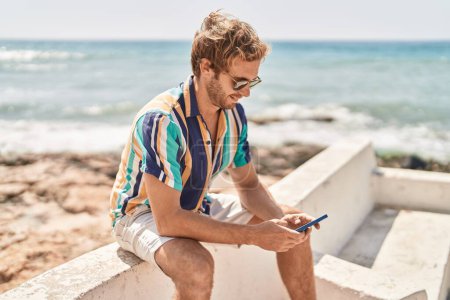 Téléchargez les photos : Jeune touriste homme utilisant smartphone assis sur le banc au bord de la mer - en image libre de droit