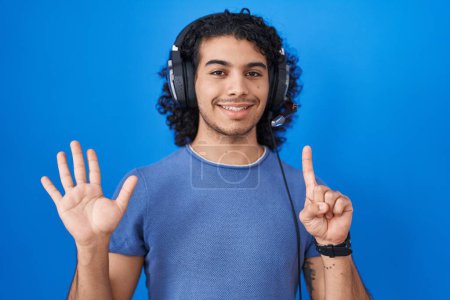 Téléchargez les photos : Homme hispanique aux cheveux bouclés écoutant de la musique à l'aide d'écouteurs montrant et pointant vers le haut avec les doigts numéro six tout en souriant confiant et heureux. - en image libre de droit