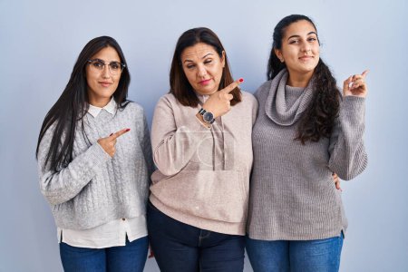 Photo for Mother and two daughters standing over blue background pointing with hand finger to the side showing advertisement, serious and calm face - Royalty Free Image