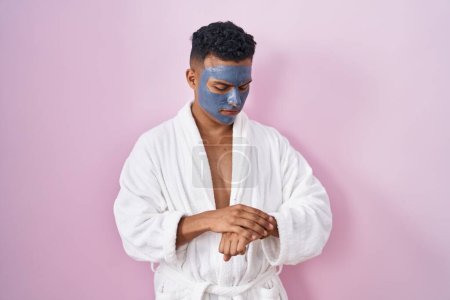 Photo for Young hispanic man wearing beauty face mask and bath robe checking the time on wrist watch, relaxed and confident - Royalty Free Image