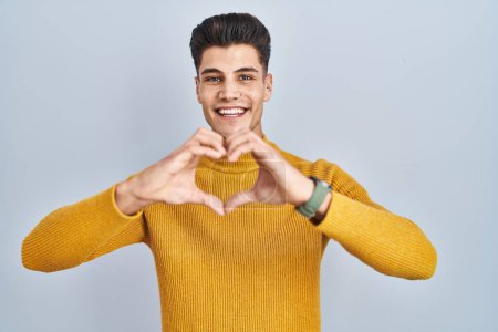 Foto de Joven hombre hispano de pie sobre fondo azul sonriendo en el amor haciendo forma de símbolo del corazón con las manos. concepto romántico. - Imagen libre de derechos