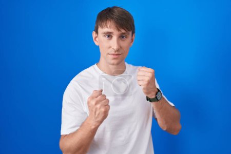 Photo for Caucasian blond man standing over blue background ready to fight with fist defense gesture, angry and upset face, afraid of problem - Royalty Free Image