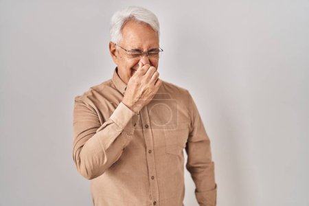 Téléchargez les photos : Hispanic senior man wearing glasses smelling something stinky and disgusting, intolerable smell, holding breath with fingers on nose. bad smell - en image libre de droit