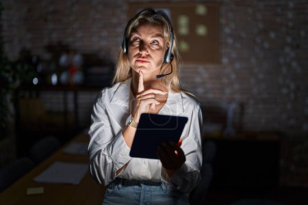 Photo for Young blonde woman working at the office at night thinking concentrated about doubt with finger on chin and looking up wondering - Royalty Free Image