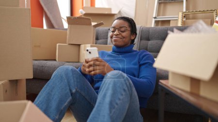 Foto de Mujer afroamericana usando teléfono inteligente sentado en el piso en un nuevo hogar - Imagen libre de derechos
