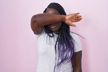 Téléchargez les photos : Jeune femme africaine debout sur fond rose couvrant les yeux avec le bras, l'air grave et triste. concept de non-visibilité, de dissimulation et de rejet - en image libre de droit