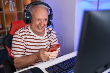Foto de Streamer de hombre de pelo gris de mediana edad usando computadora y teléfono inteligente en la sala de juegos - Imagen libre de derechos