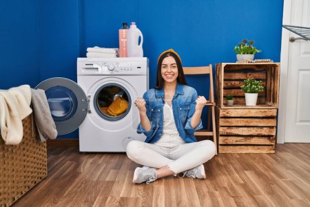 Photo for Hispanic woman doing laundry sitting on the floor screaming proud, celebrating victory and success very excited with raised arms - Royalty Free Image