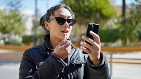 Téléchargez les photos : Jeune belle femme hispanique utilisant smartphone comme miroir maquillage lèvres au parc - en image libre de droit