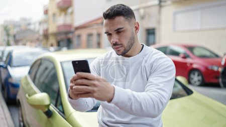 Téléchargez les photos : Young hispanic man using smartphone sitting on car at street - en image libre de droit
