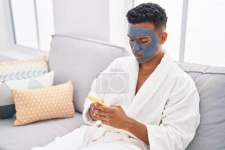 Téléchargez les photos : Jeune homme latino détendu sur canapé avec masque facial à l'aide d'un smartphone à la maison - en image libre de droit