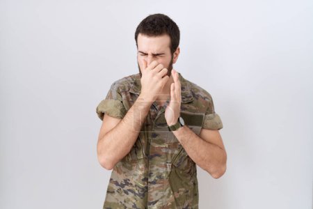Téléchargez les photos : Jeune homme hispanique portant l'uniforme de camouflage de l'armée sentant quelque chose puant et dégoûtant, odeur intolérable, retenant la respiration avec les doigts sur le nez. mauvaise odeur - en image libre de droit