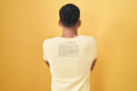 Foto de Hombre hispano con barba de pie sobre fondo amarillo de pie hacia atrás mirando hacia otro lado con los brazos cruzados - Imagen libre de derechos