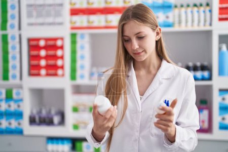 Téléchargez les photos : Jeune pharmacienne caucasienne souriante confiante tenant des bouteilles de pilules à la pharmacie - en image libre de droit