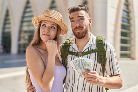 Photo for Young tourist couple holding dollars banknotes serious face thinking about question with hand on chin, thoughtful about confusing idea - Royalty Free Image