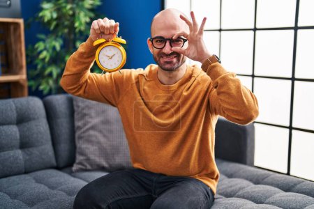 Photo for Young bald man with beard holding alarm clock smiling happy doing ok sign with hand on eye looking through fingers - Royalty Free Image